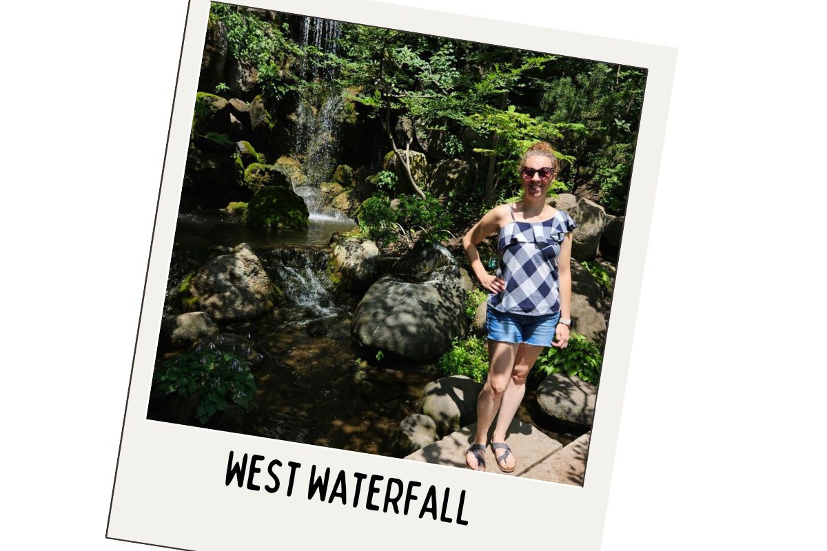 Angela is wearing sunglasses, a checkered sleeveless top, and denim shorts and is standing on a path next to a rocky waterfall surrounded by lush greenery. The polaroid image has a white border with the text "WEST WATERFALL" at the bottom.