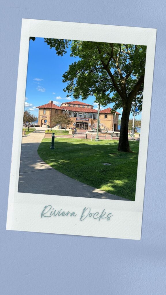 A photo of the Riviera Docks building in Lake Geneva, WI, framed by a large tree and greenery in the foreground. The path leading to the building is surrounded by well-maintained lawns, and the building itself features a classic design with red-tiled roofs and an arched entrance. The sign "The Riviera" is prominently displayed above the entrance.