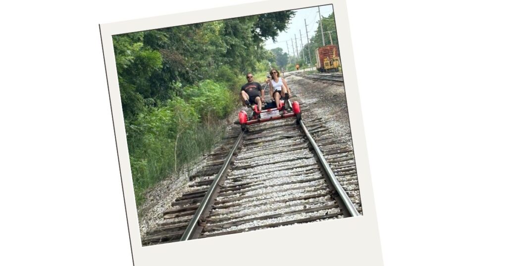 Dominic and I are riding down the railroad track on the rail bike. There are green trees on our right and another set of railroad tracks on our left. 