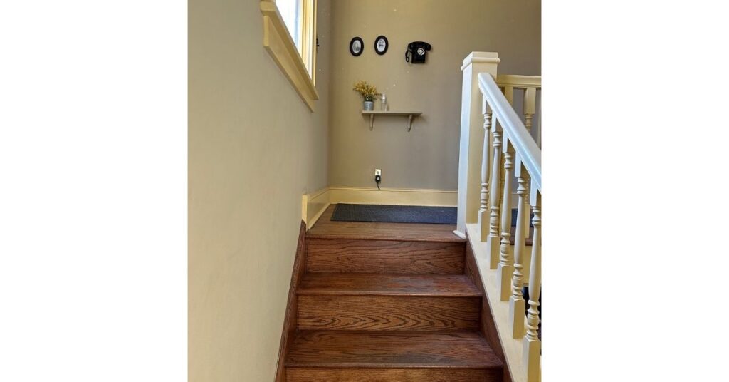 Wooden staircase with white railings. There is a small shelf with flowers, a couple of photos, and a black phone hanging on the wall at the top of the stairs.