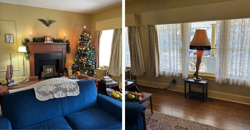 A Christmas Story house- living room. The image shows a cozy living room decorated for the holidays. On the left side, there's a Christmas tree adorned with colorful ornaments, next to a fireplace with garland and candles on the mantel. A blue couch with a white lace throw is positioned in front of the fireplace. On the right side, a window is partially covered with lace curtains, and in front of it is a distinctive lamp shaped like a leg wearing a high heel, referencing the iconic leg lamp from the movie *A Christmas Story*. The room has a warm, nostalgic ambiance with vintage furniture and decor.