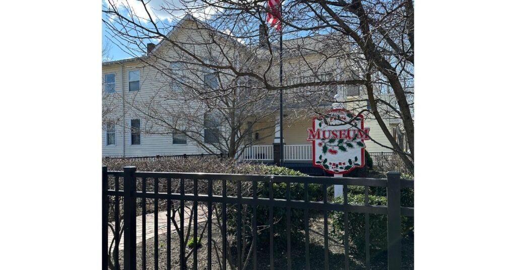 Black fence in the front of the picture with a tree behind it. In the background is a large white building. There is also a sign that says, "Museum" in red letters.