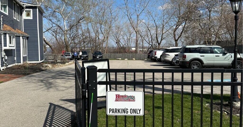 Parking lot at A Christmas Story house. There is a black fence with a sign that says, "House from A Christmas Story Parking Only". There are cars in the parking lot and the blue Bumpus House on the left.