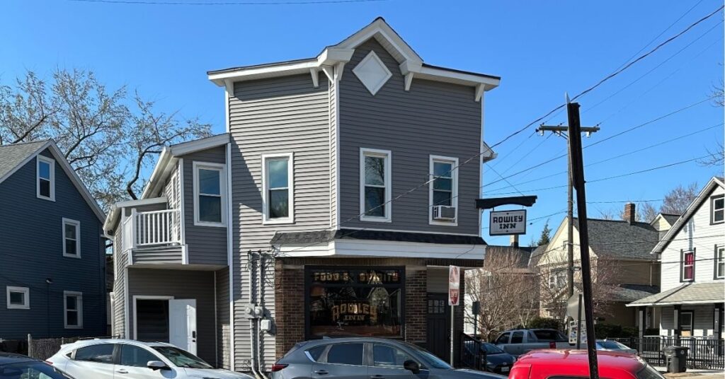 This gray building with white windows has a sign hanging from the side of it and writing on the front window that says, " Rowley Inn". There are cars parked in front of the building.