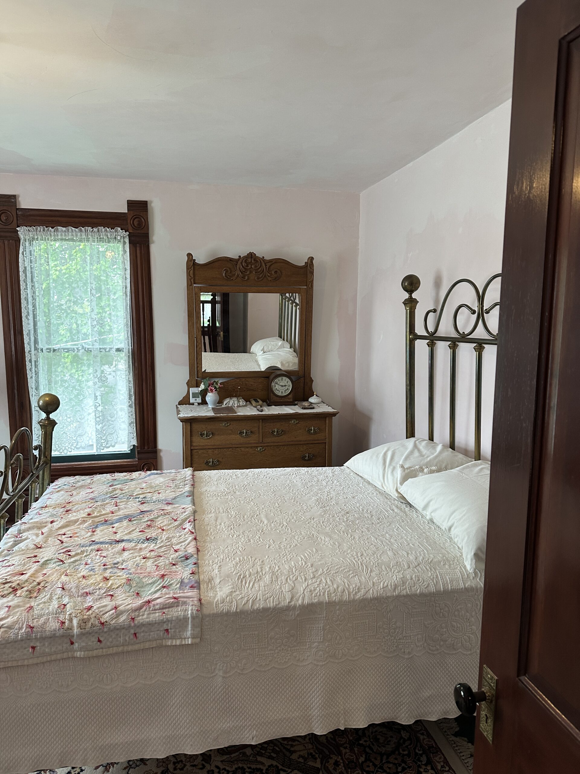 Master bedroom that has an iron headboard and white blanket on the bed. It has a second blanket with red threads spotted all over it. There is a window with lace curtains and a chest with a mirror above it in the background.