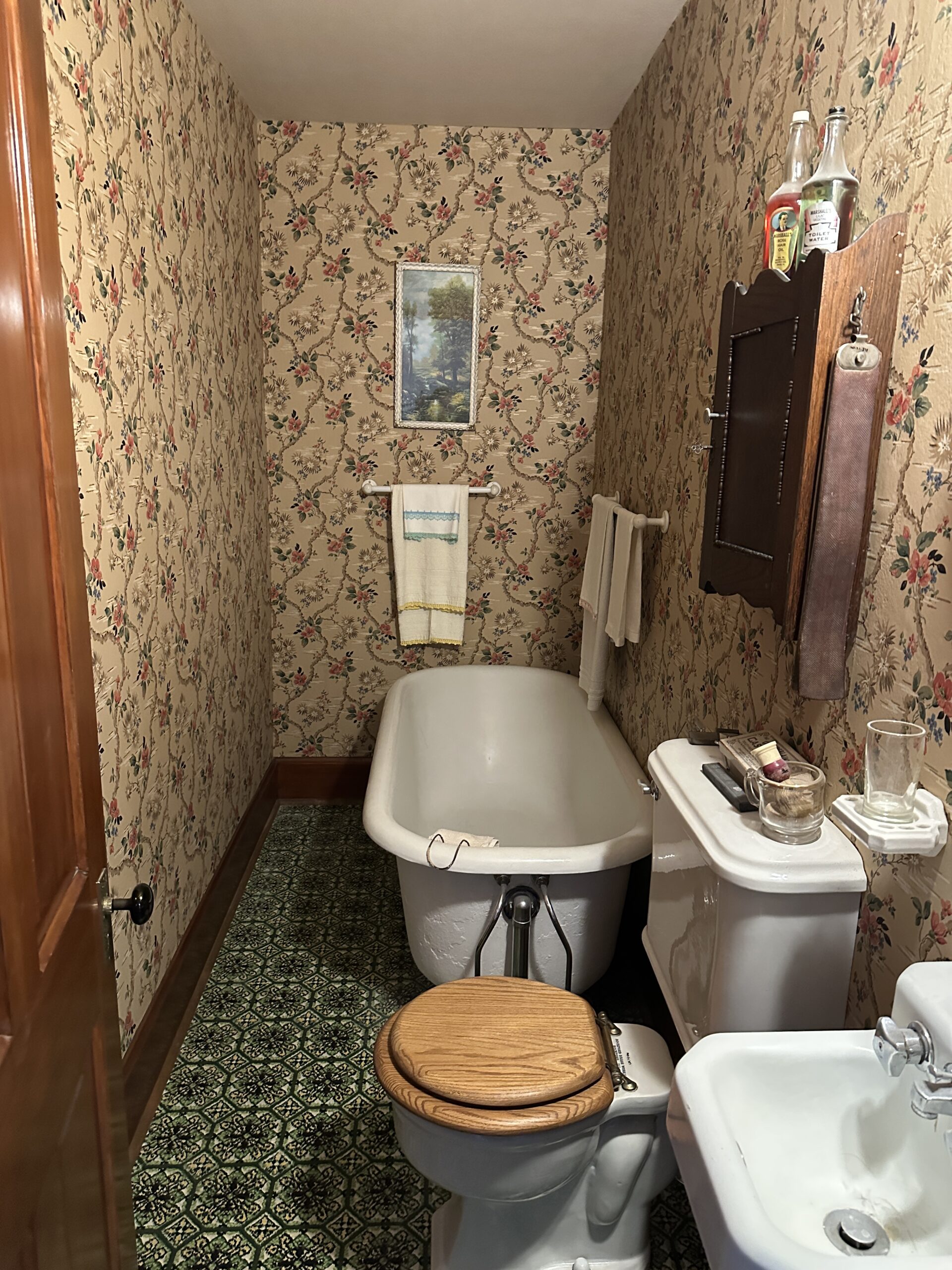 Bathroom in Reagan's boyhood home. There is a white stand alone bathtub, toilet, and white sink. The bathroom floor has green tile and the walls are covered with floral wallpaper.