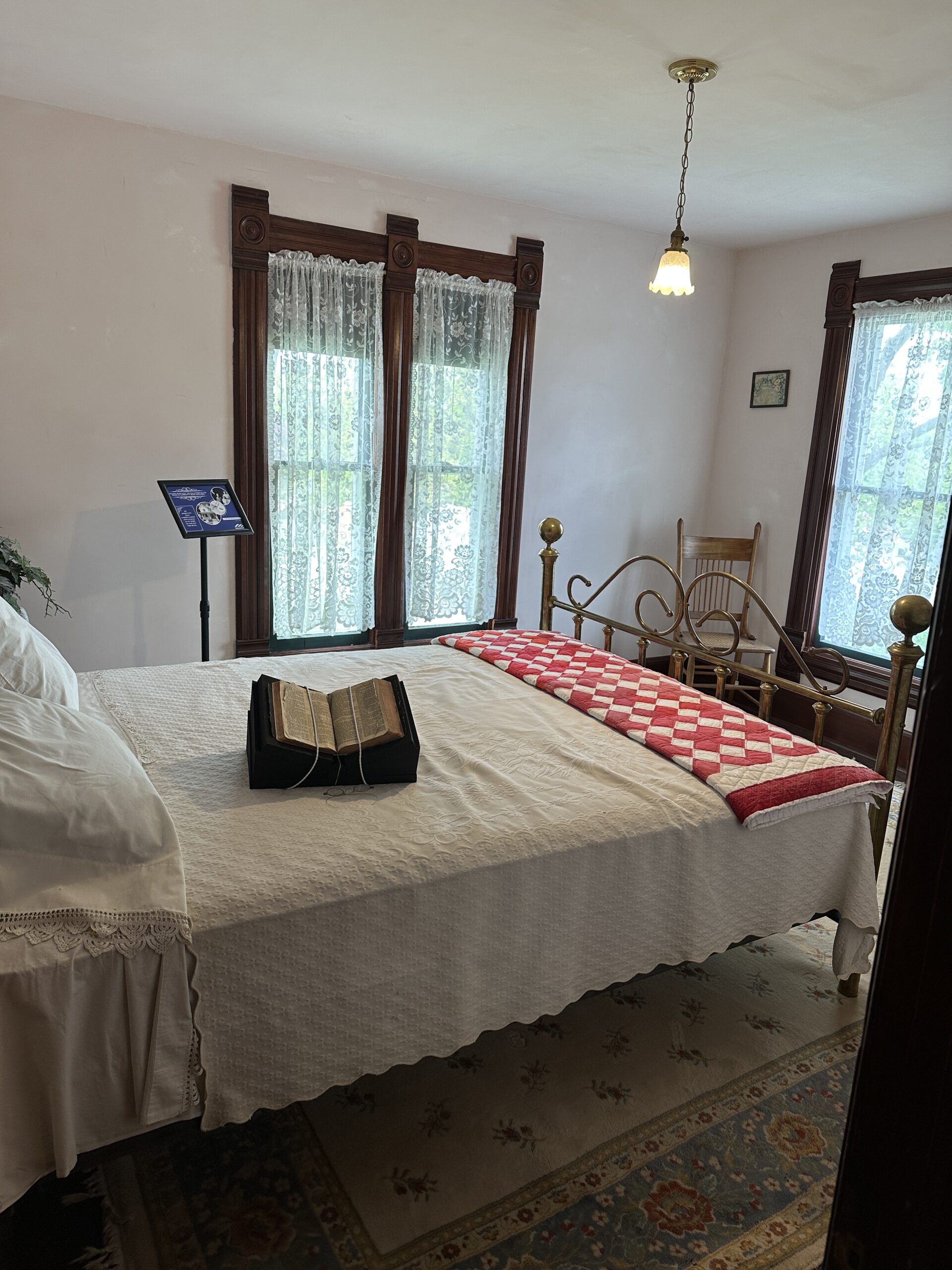 This is a picture of a bed in the guest room. There are 3 windows with white lace curtains. The bed has a white quilt with another folded red and white quilt on the foot of the bed. There is an open bible on the bed..