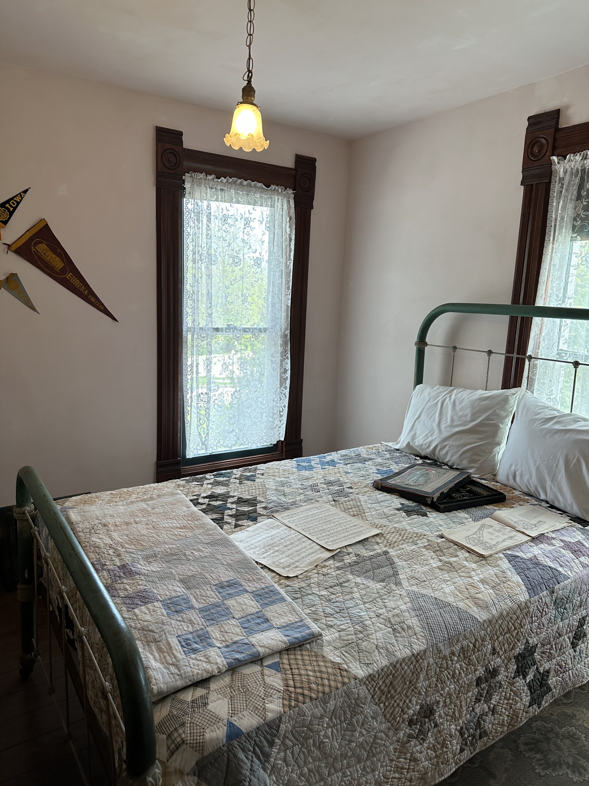 This is a picture of Ronald Reagan and his brother's bedroom that they shared. There is a patterned quilt on the bed along with some papers. The windows has lace curtains on them and there are some pennants on the white wall.