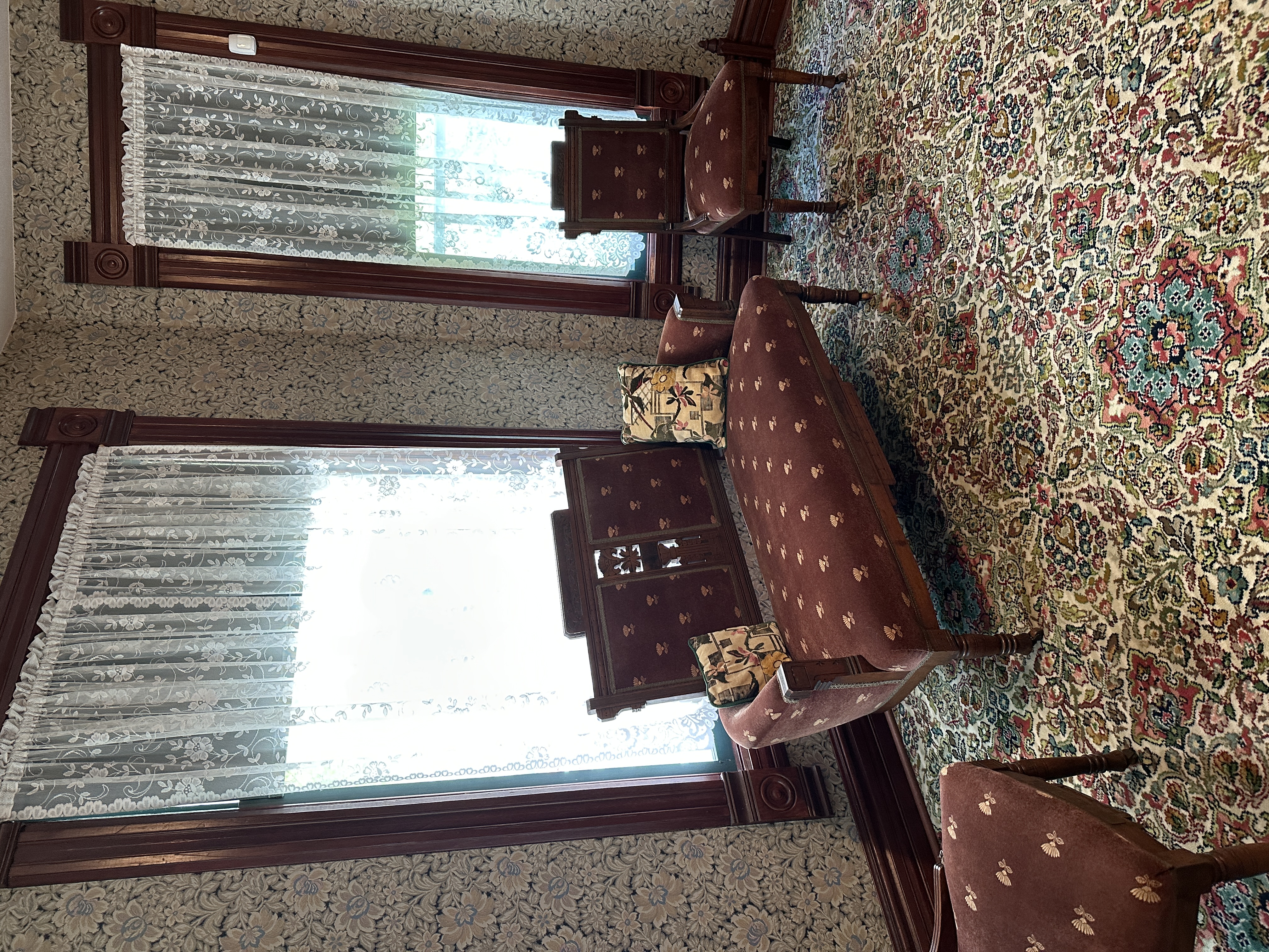 Victorian-style sitting room in Reagan's boyhood home with ornate floral wallpaper, lace curtains, and carved wooden window frames. The room features a vintage upholstered settee and matching chairs in burgundy fabric with gold motifs, set on a richly patterned Persian-style carpet.