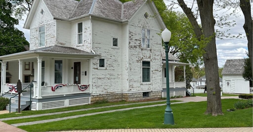 Ronald Reagan's boyhood home in Dixon, Illinois—a two-story white wooden house with peeling paint, steep gabled rooflines, and a covered front porch decorated with red, white, and blue bunting. The house is set on a neatly maintained lawn with stone walkways and mature trees. A green lamppost stands in the foreground, and a small white outbuilding is visible in the background under a partly cloudy sky.
