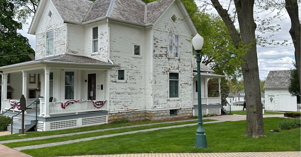 Ronald Reagan's boyhood home in Dixon, Illinois—a two-story white wooden house with peeling paint, steep gabled rooflines, and a covered front porch decorated with red, white, and blue bunting. The house is set on a neatly maintained lawn with stone walkways and mature trees. A green lamppost stands in the foreground, and a small white outbuilding is visible in the background under a partly cloudy sky.